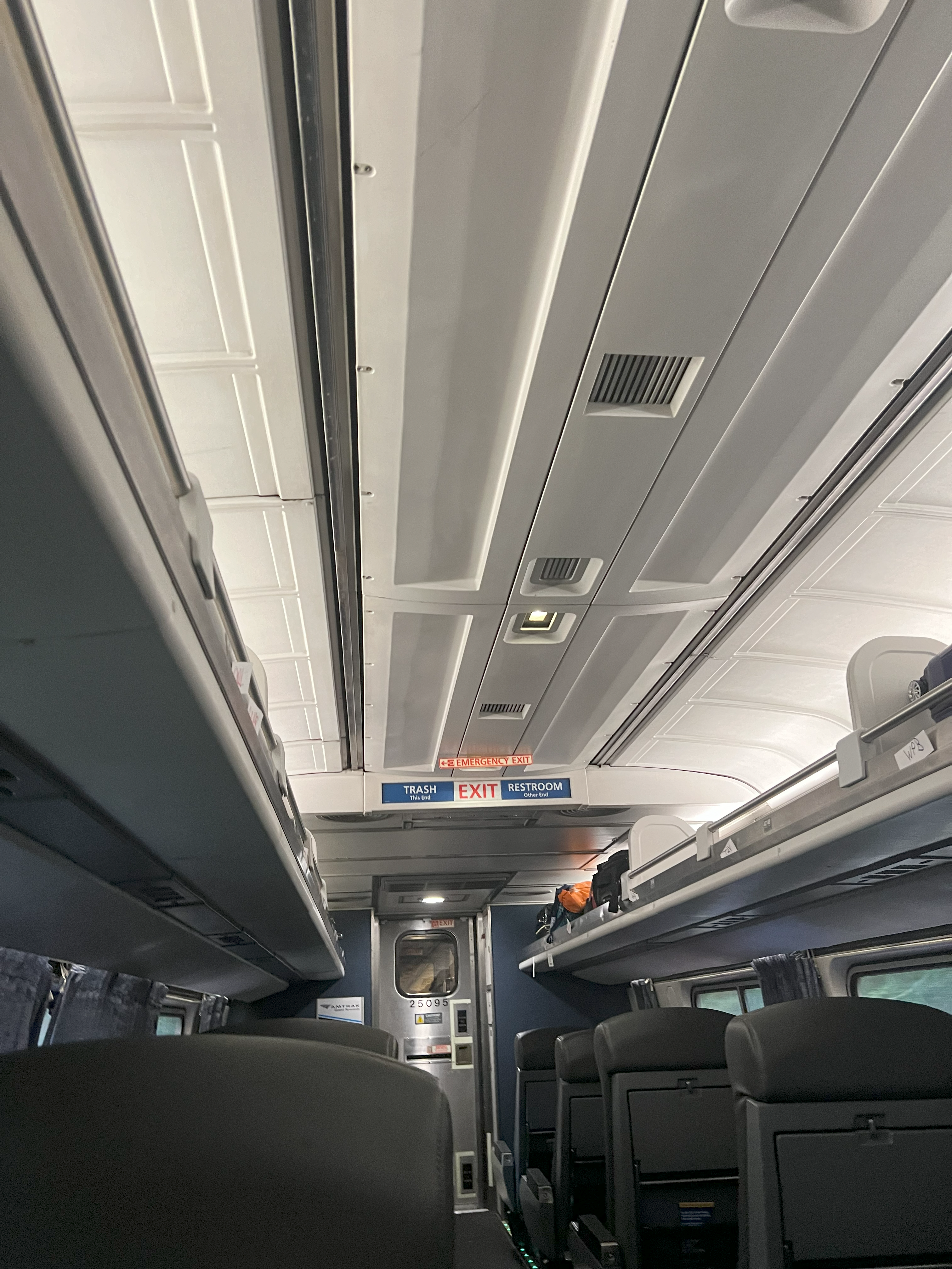 Interior view of an Amtrak train coach showing overhead luggage racks, passenger seats, and exit/restroom signage